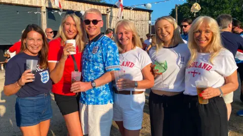 Angela Ferguson/BBC A group of five women and one man are smiling and holding drinks while standing outside The Big Barn.
