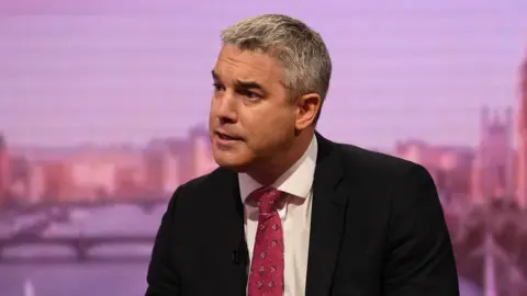 Sir Stephen Barclay being interviewed in a studio. He is wearing a dark-coloured jacket, white shirt and red tie. 