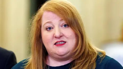 Liam McBurney/PA Wire Naomi Long has auburn hair. She is wearing a silver necklace and a top with black trousers. Someone out of shot is standing next to her.