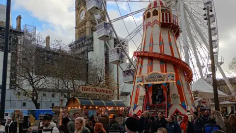 Crowds flock around a helter-skelter and a Ferris wheel by a hot chestnuts stall in Albert Square