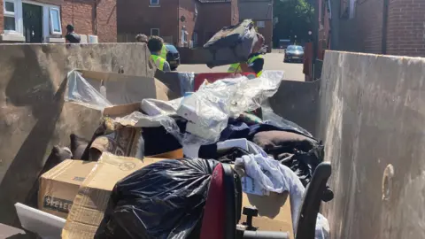 OLI CONSTABLE/BBC A close-up shot from the rear of a skip. Rubbish is inside it including a chair, a carpet and cardboard. A man can be seen putting items into it.