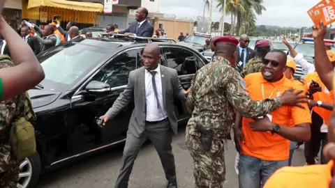 Egnan Koula/EPA/Shutterstock Ivorian President Alassane Ouattara stands up through the roof of a sunroof of a black car surrounded by security officers and his supporters as he arrives at the electoral commission in Abidjan - Tuesday 26 August 2025. 