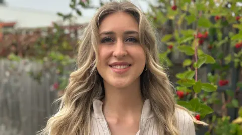 Handout A woman with long blonde hair smiling at the camera. She is wearing a light coloured top and standing in front of a wooden fence with trailing plants and red flowers growing in front.