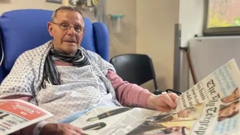 Hospital patient Alfred Mayes sits in a hospital chair reading a newspaper.