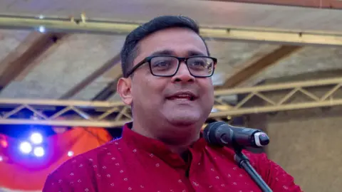 Chirag Khajuria with short black hair wearing a red sherwani on stage talking into a microphone.
