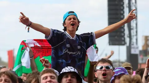 A boy, wearing a blue cap and blue T-shirt, being lofted above on a person's shoulders, who is spreading his arms out wide as he sings to a song at the festival. 