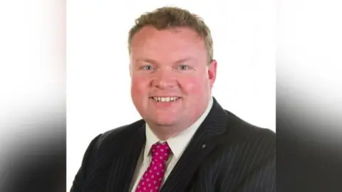 Whiteabbey Primary School Keith Wysner smiling wearing a pinstripe blazer, a white shirt and a red patterned tie.