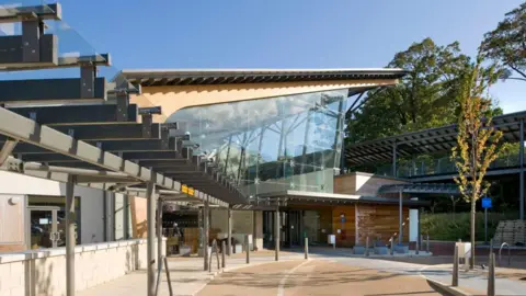 The entrance to a hospital with a road and path leading up to it. The main building has a slanted ceiling and a large open window either side with two longer buildings either side of it.