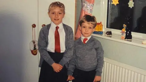 The childhood photo of Claire and Peter which was sent to Dalibor Sentic in the care package he recieved in 1992. Claire and Peter are photographed in school uniforms, stood in what appears to be a bedroom. Claire is taller than Peter and stood to the left of the image. She is wearing a black skirt, white blouse, red tie, and grey cardigan. She has blonde hair with a fringe. Her hair is tied up. Peter is shorter than Claire and is photographed to the right of her. He is wearing black trousers, a white shirt and red tie, with a grey jumper over the top.