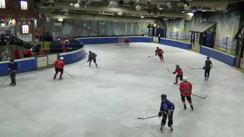 A number of ice hockey players playing on an undercover ice rink. They are all wearing helmets and carrying hockey sticks. They are split into a red team and a blue team.