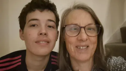 Melody Pilgrim A woman with brown hair and glasses sat on a staircase with a white wall. Also in the close-up selfie is her teenage son, who has black hair and is wearing a black sports top. Both people are smiling.