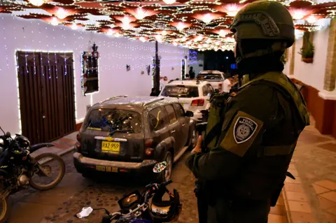 AFP via Getty Images An officer with an arm patch saying 'policia nacional' looks on while dressed in military gear as hearses carrying bodies of the victims drive down a street in Playa de Belen on 28 January