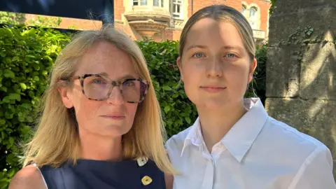 A woman wearing a dark blue dress is standing next to a girl wearing a white shirt. They are next to a hedge and a stone wall. Further behind them is a red brick building with stone window surrounds and elaborate bay windows.