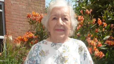 Phyll Babb Phyllis Babb. She has short white hair and is pictured outside, in front of some orange flowers. She is wearing a white top with a floral pattern and a silver necklace. She is looking directly at the camera and smiling.