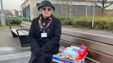 Vera Parnaby, sitting on a wooden bench next to a box of paper poppies and a collection tin. She is wearing a black hat, coat and gloves as well as shaded glasses and a lanyard containing her seller information.