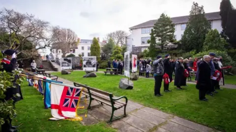 Royal Guernsey Light Infantry Trust People stood in a park during a military memorial service. A group of people on the left-hand side are holding flags on poles which have been lowered to the ground. Other people stood on a patch of grass have poppy wreaths in their hands.
