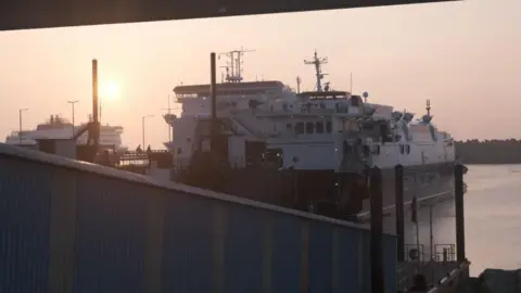 Isle of Man Government A view of the rear of Isle of Man Steam Packet fast craft Manannan moored in Douglas Harbour with a ramp down to the vessel in the foreground as the sun sets in the background, where another ferry can be seen in the distance. 