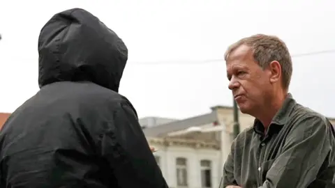 Man in back coat with hood up faces Andrew Harding
