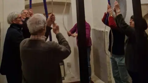 Joe Wheller A group of people pulling on ropes to ring bells. They are stood in a circle.