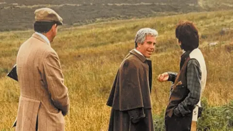 US Department of Justice Jeffrey Epstein, wearing what appears to be a cape in brown wool or tweed, turning towards a woman with short brown hair, who resembles Ghislaine Maxwell. To the left is a man in a light brown tweed jacket and a flat cap, with the stock of a shotgun tucked under his arm. Behind them is a hilly landscape with bracken and dry grass. 