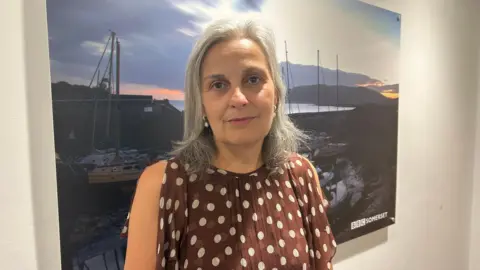 A woman with shoulder length grey hair stands and looks at the camera with a neutral expression. She is wearing a brown dress with white dots. Behind her is a photograph of a harbour.