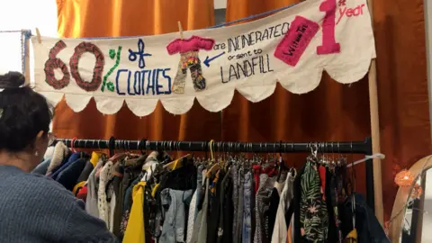 Molly Smith / BBC A clothes rail with lots of different types of garments hung on it. There is some bunting above it with words embroidered on it, saying: 60% of all clothes are incinerated or sent to landfill within the 1st year. There is a woman browsing the clothes and she can be seen from behind.