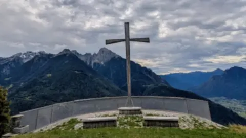 Submitted A large cross on top of a raised platform has a grey barrier in front of it. It overlooks several mountain tops on a cloudy day.