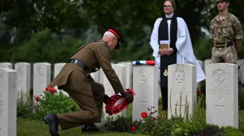Crown Copyright A uniformed service member kneels to lay a red poppy wreath at a row of Commonwealth war graves.