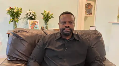 A man with short black hair and a goatee sits on a brown, leather-look armchair. Behind him is a framed photograph of a young man, his son, smiling with his thumbs up in a pool. There are three vases of flowers around the photo.
