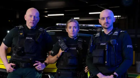 BBC/ Third Street Studios Three police officers looking at the camera. All are wearing police uniform that is black and dark green. The one on the left is a bald man, in the middle is a blonde woman and on the right is a bald man with dark beard.