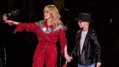 A woman in a red outfit with a boy wearing a black leather jacket, white t-shirt and black hat