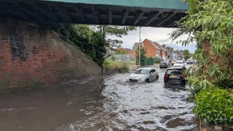 A silver Mercedes-type car is being driven into deep water, underneath a railway bridge. Another car, which is dark in colour, is being driven away from the camera through the water. There are terraced or semi-detached houses in the background.