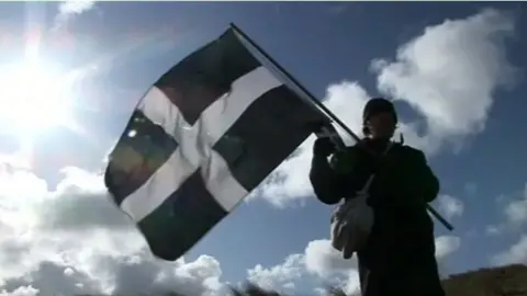 BBC A silhouetted figure holding a Cornish flag against a blue sky