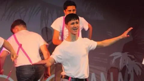 Conor is dancing on stage, wearing a white shirt, belt and jeans, with dangling pink braces.