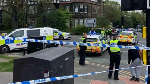 Cathy Killick/BBC A police cordon in place at a busy junction. Present are several uniformed officers standing on the street corner, with four police vehicles around them. A roadsign, pedestrian crossing and other street furniture can be seen.