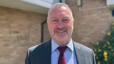 Steve Reed, a man, with short grey hair, wearing a grey suit and a red tie with a blue checked shirt, smiling for camera.