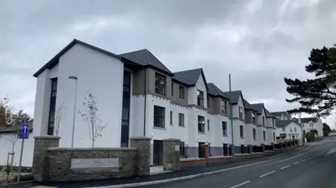 The exterior of the Mayfield Apartments, which is a large white and brown building with a road running alongside it.