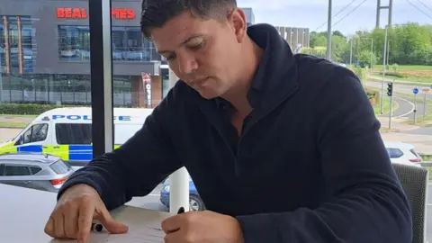 Luke Campbell with short, dark hair wearing a blue top signing the acceptance of office declaration in an glass sided office with a view of the Humber Bridge in the background