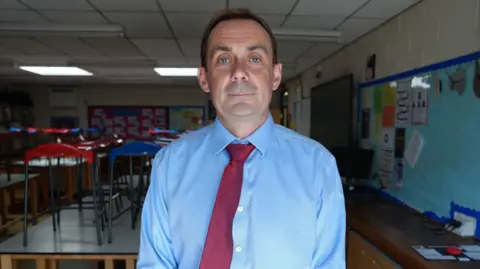 Qays Najm/BBC A man stands in a classroom. He has short dark hair and wears a light blue shirt and red tie. In the classroom the chairs sit on the tables.