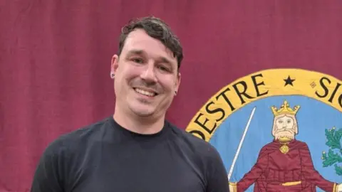 A man with dark hair and a dark tshirt in front of an embroidered picture of a king in red robes