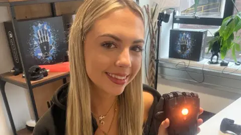 Sadie, who has long blonde hair and is wearing grey tracksuit bottoms, a black top and a black jumper, is holding up the prosthetic hand, which has an orange light flashing from the centre of it.
