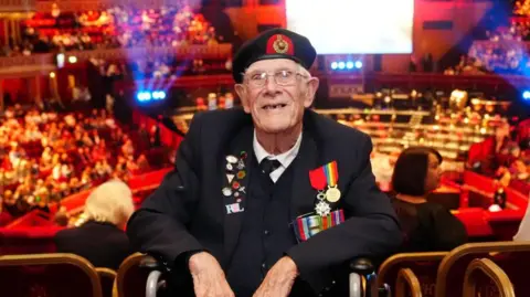 Jim Grant during a concert celebrating the 80th Anniversary of VE Day. He is sitting in a wheelchair and is wearing his veteran uniform. Behind him is the Royal Albert Hall, with an audience and lights.