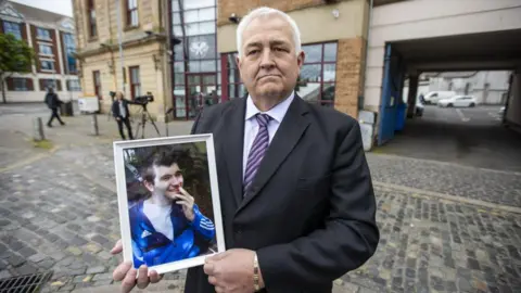 Glynn Brown has short white hair. He is wearing a lilac shirt, purple tie and black suit, holding a framed photo of a young man, who is wearing a blue tracksuit top over a T-shirt. He has a hand raised to his face and is smiling.