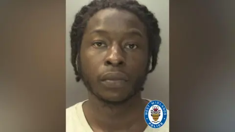 A mugshot of Elie Mey wearing a white t shirt. He has black braided hair and has a short black beard