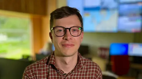 Martin Giles/BBC Fred Best is wearing dark-rimmed glasses and has short brown hair and is wearing a red checked shirt. He is sitting down