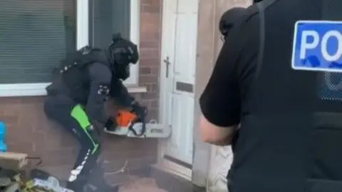 A police officer wearing protective clothing uses a chainsaw to cut through the front door of a house, while another officer stands watching.