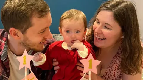 A man, a woman and a baby sit behind a cake with a candle in it