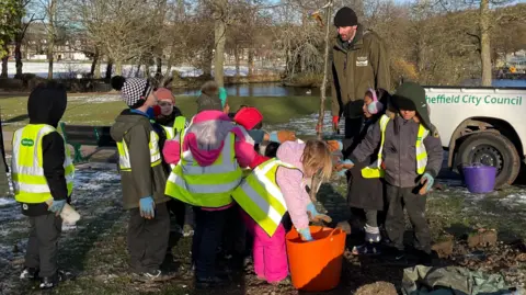 TOM MACDOUGALL/BBC The children are gathered around the sapling, listening to a man from the council who's talking. One girl is reaching into an orange bucket filled with food for the tree.