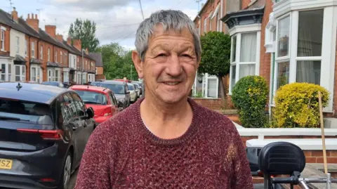 Man with short hair and a dark red jumper. he is standing on a residential street with cars parked along it.