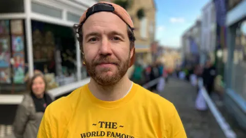 A man in a yellow T-Shirt is standing on a cobbled street, looking at the camera. He is wearing a cap backwards and is smiling. There are people in the background lining the street.